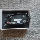 Černý snímač s připojenými vodiči v bílé krabičce s pěnovou výplní. Na spodní straně je nápis DIMARZIO PICKUPS a pod ním písmeno M. Vedle nápisu je n…