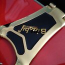 Detail zlatého vibrační mechanismu na červeném povrchu nástroje. Na černém plastovém prvku je zlatý nápis „Bigsby“ a nad ním menší text „LICENSED“. P…