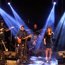 Fotografie zachycuje hudební kapelu na pódiu s osvětlením. Vzniká zde sestava tvořená baskytarou, elektrickou kytarou, bicími, saxofonem a zpěvačkou…