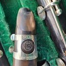 Detailní pohled na mouthpiece klarnetu s kovým ligaturem. Na ligaturem je vyražen nápis SELMER a níže kulatý znak s textem HENRI SELMER PARIS. V doln…