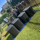 Pět černých subwooferů v hranatých bednách s kovovými mřížkami, umístěných vedle sebe na modré plachtě na trávníku. Povrch beden vykazuje drobné oděr…