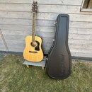 Akustická kytara s přirozenou dřevěnou barvou korpusu a tmavým hmatníkem. Na hlavě je viditelné logo Tanglewood. Vedle kytary stojí černý tvrdý kufýr…