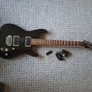 Černá elektrická kytara s tmavým hnedým hmatníkem a dvěma humbuckery. Na hlavě nástroje je bílý nápis Ibanez. Vedle kytary leží černý napájecí adapté…