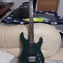 Tmavě zelená elektrická kytara s dvojsekmovým tělem a hnědým hmatníkem. Na hlavě je viditelné zlaté logo Kramer. Nástroj je vybaven dvěma humbuckery…