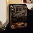 Detail chromovaného krytu elektroniky na černé kytáře. Uprostřed je vyryto logo Squier by Fender. Kryt je připevněn čtyřmi šrouby a vykazuje mírné zn…
