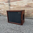 Hnědý basový reproduktor v dřevěném korpusu s černou textilní maskou. V pravém horním rohu masky je umístěn štítek s nápisem „gbr Custom Cabinets“. P…