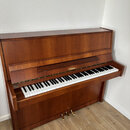 Hnědé dřevěné piano s otevřenou klaviaturou. Na přihradě nad klávesami je zlatý nápis „Behringer“. Nástroj vykazuje známky běžného používání, povrch…