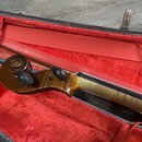 Detailní pohled na hmatník a hlavu smyčcového nástroje v červeně vystlaném pouzdře. Nástroj je vyroben z lakovaného dřeva s viditnou kresbou zrna na…