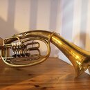 Mosazný dechový nástroj s nápisem B na trubici. Nástroj má čtyři ventily s bílými knoflíky a viditelné stopy opotřebení povrchu v podobě drobných škr…