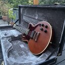 Elektrická kytara s hnědým trupem a tmavě červeným pickguardem v černém kufru s šedým polstrováním. Nástroj má dva humbuckery, tři oválné černé knofl…