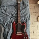 Elektrická kytara s červeně hnědým lakem a černým pickguardem. Na hlavě krku je viditelné logo Epiphone. Nástroj vykazuje mírné opotřebení povrchu v…
