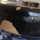Detailní záběr na černý lakovaný povrch hudebního nástroje. Na hraně jsou vidět dvě výrazná odloupení laku, pod kterým je světle hnědé dřevo s vidite…
