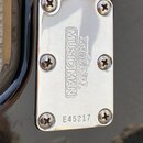 Detail chromovaného krycího krytu na černém povrchu nástroje. Na krytu je vyražen nápis ERNIE BALL MUSIC MAN a sériové číslo E45217. Povrch krytu vyk…