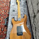 Zadní strana elektrické kytary s přírodním dřevěným povrchem v tvrdém kufru. Na krku je viditelný štítek s nápisy „Custom Shop“, „Fender“ a nečitelný…