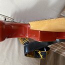 Detailní pohled na spoj krku a trupu červené elektrické kytary. Krk je vyroben ze světlého dřeva. Pod nástrojem je vidět černý popruh s žlutými prvky…