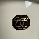 Detailní záběr na bílý, lesklý povrch s osmiúhelníkovým černým štítkem. Na štítku je zlatým písmem nápis Gretsch a pod ním text DRUM MAKERS SINCE 188…