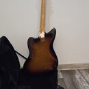Elektrická kytara s trupem v barevném přechodu od žluté přes hnědou k černé (sunburst). Na hlavě krku je viditelné logo "Fender". Kytara je opřena o…