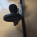 Černý mikrofon s pěnovou windscreenovou krytkou a černým popfiltrem. Na popfiltru je bílý nápis RØDE. Soubor doplňuje černý mikrofonní stojan z kovu.