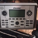 Stříbrošedý modul elektronických bicích s černým displejem a ovládacími prvky. Na horní části je logo CARLSBRO a v dolní části nápis COMMANDER 500. M…