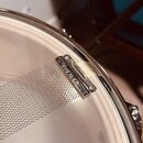 Detailní pohled na kovový snare wire (struny) u bubnu. Na kovovém držáku je vyryt nápis PREMIER. Povrch kovu vykazuje známky mírného opotřebení a oxi…
