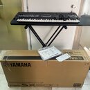Černý digitální klavír umístěný na černém X-stojanu. Pod nástrojem je hnědý kartonový obal s nápisy YAMAHA, PSR-SX600 a DIGITAL KEYBOARD / CLAVIER NU…