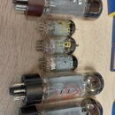 Fotografie zobrazuje skupinu elektronických vakuových elektronek různých velikostí a typů položených na dřevěném povrchu. Na jedné z elektronek je či…