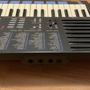 Šedý digitální klavír značky YAMAHA s bílými a černými klávesami. Pod klaviatura je panel s ovládacími prvky a tabulkou s názvy hlasů pod nadpisem VO…