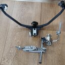 Fotografie zobrazuje dvě části kovového hardware pro bicí nástroje v černé a chromované barvě. Horní část tvoří černý oblouk s dvěma uchopovadly na k…