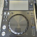 Černý DJ player značky Pioneer DJ, model MULTI PLAYER XDJ-700. Zařízení obsahuje displej, velký ovládací jog s vnitřním zrcadlovým povrchem a vertiká…