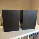 Dva černé reproduktory s povrchem imitujícím strukturu dřeva. Jsou umístěny vedle sebe na bílém nábytku. Na přístrojích nejsou viditelná žádná loga a…