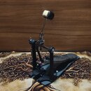 Černý basový pedál s kovovou konstrukcí a stříbrnými mechanickými prvky. Na šlapadle je vidět vyryté logo, které je však nečitelné. Povrch nástroje v…