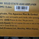 Fotografie detailu štítku na zlatavém kovovém povrchu. Na štítku je text MODEL 8080 SOLID STATE LEAD AMPLIFIER, 80 Watts RMS / 8 ohm a čárcový kód s…
