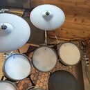 Elektronická bicí souprava s bílými pady a činely. Na povrchu padů a činelů je viditelné logo značky Roland. Souprava je umístěna na černém stojanu n…