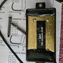 Černý humbucker s mosazným krytem a černým štítkem, na kterém je bílý nápis RMC RM PICKUPS 5/26. Snímač má v horní a spodní části montážní uši s otvo…