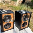 Dva černé hranaté reproduktory s odobračkami v oranžovém odstínu. Každý box obsahuje jeden větší a jeden menší kruhový reproduktor; u obou je viditel…