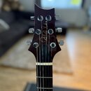 Fotografie zachycuje hlavu a horní část hmatníku elektrické kytary v tmavě vínové barvě. Na hlavě je bílý nápis "Paul Reed Smith". Nástroj má šest st…