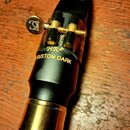 Černý saxofonový mouthpiece s mosazným prstencem u patky. Na povrchu je zlatým písmem nápis HR* CUSTOM DARK a nad ním nečitelný podpis. Mundstuk je v…
