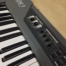 Šedý syntetizátor s klávesnicí. Na horním panelu je nápis JX-1 SYNTHESIZER a ovládací prvky s popisky VOLUME, BRILLIANCE, MELLOW, BRIGHT a sekce EDIT…