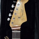Fotografie zachycuje hlavu a část hmatníku elektrické kytary v černém obalu. Na zadní straně světlé dřevěné hlavy je nápis Fender a JAZZMASTER, dále…