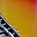 Detailní záběr na část těla kytary s žlutě-oranžovým přechodem barev (sunburst). V levé části jsou vidět kovové sedla kobylníku. Povrch laku vykazuje…