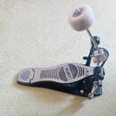 Černý basový pedál s kovovou stoupací plochou. Na pedálu je vyražen nápis MAPEX. Horní část mechanismu obsahuje bílé plastové kolečko na kovovém rame…