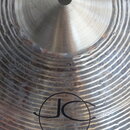 Detailní pohled na kovový činel s výrazným rýhováním povrchu. Na spodní části je černé logo „JC“ v kruhu a nápis „14" Sparkle Hi-Hat Bottom“. Povrch…