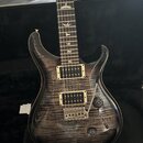 Elektrická kytara s tmavým, žilnatým dřevěným povrchem korpusu a hnedým hmatníkem. Nástroj má dva humbuckery se stříbrnými kryty, vibrační systém a d…