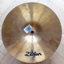 Zlatý kovový činel s viditelnými stopami používání a mírnými skvrnami na povrchu. V dolní části je modrý nápis Zildjian. U středu činelu je nalepená…