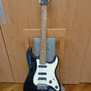 Černá elektrická kytara s bílým pickguardem a hnedým hmatníkem. Na hlavě je viditelné logo Squier. Nástroj má tři snímače typu single-coil a chromova…