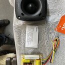 Černý reproduktor v plastovém rámu s logem JBL. Vedle něj je bílý papírek s nápisem CX380 a textem o zapojení. Dole je umístěna elektronická deska s…