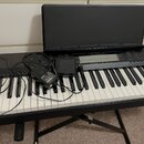 Černý digitální klavír na černém X-stánu s přiloženým černým music standem. Na ovládacím panelu je nápis MUSICAL INFORMATION SYSTEM a několik tlačíte…