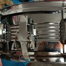 Detailní pohled na chromovaný kovový obručový tamburín. Nástroj má zrcadlový povrch s vodorovnými žebrováním a viditelným mechanismusem pro upevnění…