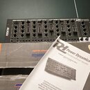 Černý mixážní pult s nápisem PDZM700 na levé straně a logem R1 Power Dynamics v pravém horním rohu. Zařízení leží na hnědých kartonových krabicích, p…