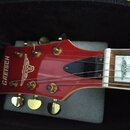 Detail hlavy červené elektrické kytary s nápisem GRETSCH a doprovodným logem. Na hmatníku je vidět bílý vložkový znak s motivem. Nástroj má zlaté lad…